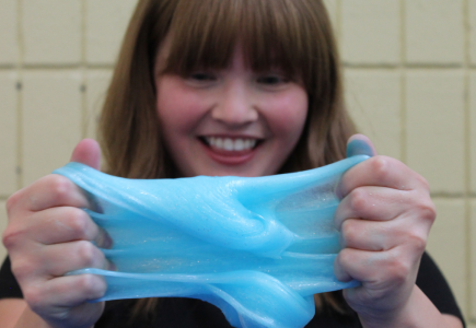 young woman smiles, blue slime is stretched in the foreground between her hands