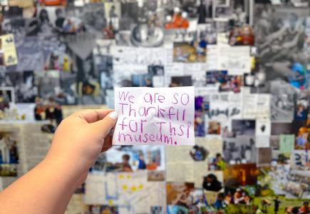 A hand holds up a note written by a child in pink crayon. The note reads: "We are so thankful for this museum"