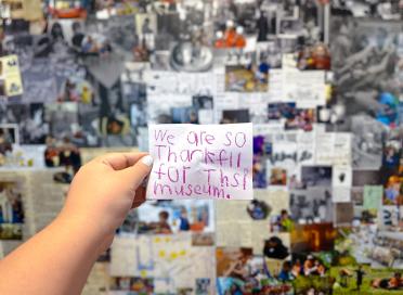 A hand holds up a note written by a child in pink crayon. The note reads: "We are so thankful for this museum"